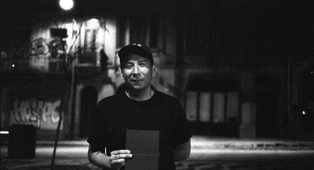 A black-and-white photograph of Sky Hopinka facing the camera on an street at night, holding a book.